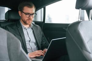 In glasses, working by using laptop. .Man in formal business clothes is sitting in the modern automobile photo