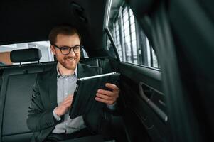 Smiling, using tablet. Man, in formal business, clothes is sitting in the modern automobile photo
