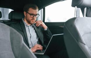 In glasses, working by using laptop. Man in ,formal business clothes is sitting in the modern automobile photo