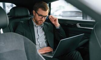 With laptop, working... Man in formal business clothes is sitting in the modern automobile photo