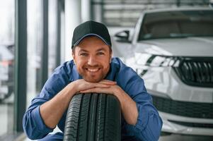 Leaning ,on, the tire and smiling. Man in blue uniform is working in the car service photo