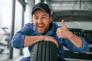 Leaning on the, tire and, smiling. Man in blue uniform is working in the car service photo