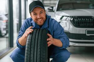 Leaning on the tire and smiling.. Man ,in ,blue uniform is working in the car service photo