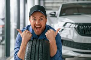 Leaning on the tire and smiling. Man in blue, uniform is working in the car service photo