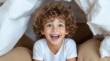 joyful child with curly hair smiling widely under cozy blanket fort photo