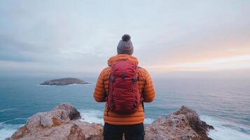 traveler in orange jacket gazes at ocean sunrise from rocky cliff photo