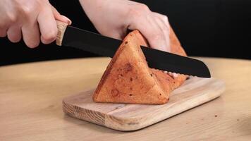Hands cutting a triangular loaf of bread with a serrated knife on a wooden board, close-up view highlights the texture and shape of the baked crust video