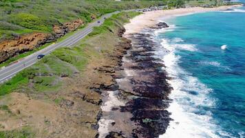 Coastal highway winds through green cliffs beside rocky Hawaiian shore, leading to a sandy beach with turquoise waves and people enjoying the view video