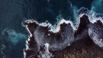 Aerial view of waves crashing against jagged black lava shoreline in Hawaii, creating dramatic white foam patterns in deep blue ocean waters video