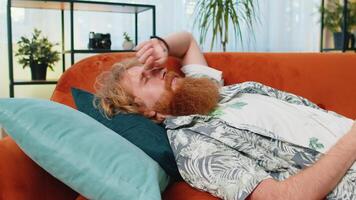 Tired Caucasian redhead man lying down in bed taking a rest at home, napping, falling asleep on sofa video