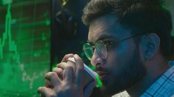Close up face Indian Arabian man holding cup drinking coffee male thoughtful guy programmer coder worker ponder analysing trader freelance trading chart home neon hacker IT specialist night solution video