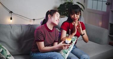 Two young women sing a happy birthday song together ,The younger sister holds a small cake with lit candles and blows out the candles on her birthday on the couch in a cozy home. video