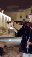 A talented girl performs composition on the instrument on the background of tall buildings in the city center. The musician plays the violin in the evening. Blurred background. Vertical video