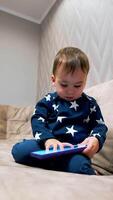 Cute toddler in blue suit is busy with phone. Kid sits on sofa focused on the smartphone and then looks at camera touching it with his finger. Low angle view. Vertical video