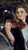 A talented girl in warm clothes is performing composition on the violin in a winter park in the evening on a blurred background. Camera motion to right Vertical video