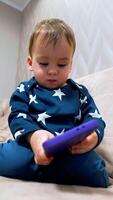 Beautiful baby boy sitting calm on sofa and looking on the phone in his hands. Lovely kid distracts on the camera and touches it with finger. Low angle view. Vertical video