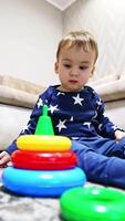Calm pretty baby boy sitting on the floor with his toys around. Cute toddler disassembles the pyramid. Low angle view. Vertical video