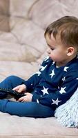 Lovely little toddler sitting cozy on the sofa. Nice kid in blue suit exploring the TV remote. Side view. Vertical video