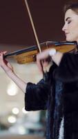 A romantic girl in warm clothes is playing a violin in the street in the square on the background of urban architecture in the evening. Blurred background. Vertical video