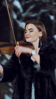 A beautiful girl with make-up and in a white dress is playing a violin composition in a winter park on the background of snow. Camera motion to left Vertical video