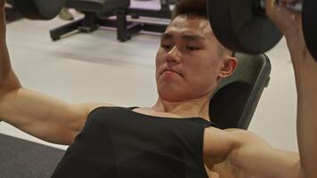 A young Asian man is working out at the gym, lying on a bench and lifting heavy dumbbells while performing the bench press. video