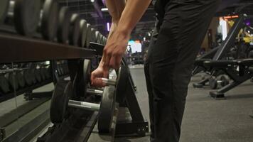 Muscular man in sweatpants stands in front of row of dumbbell racks, arranged in order of increasing weight. He selects comfortable weight to perform bicep exercises. video