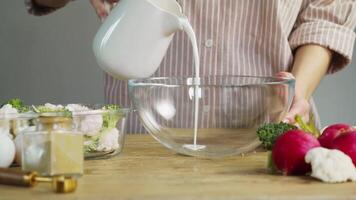 Chef in home kitchen is pouring milk or cream from saucepan into a deep dish on rustic wooden table, surrounded by fresh vegetables. video