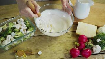 To make the sauce for the vegetable casserole with cream and cheese, the cook manually uses a wire whisk to whisk the ingredients in a bowl. video