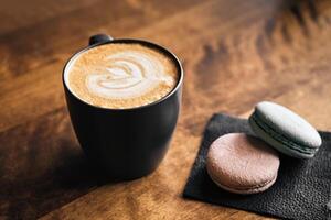 Cappuccino coffee with latte art on wooden table. Black cup, beautiful milk foam and macaroon cookies. photo