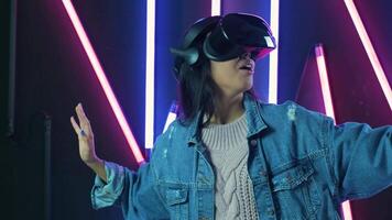 Young woman shocked, looks around and wonders how amazing in the VR headset. Virtual reality helmet on color lighting background video