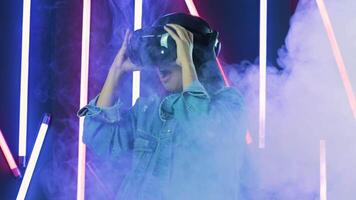 Young woman shocked, looks around and wonders how amazing in the VR headset. Virtual reality helmet on color lighting smoke background video