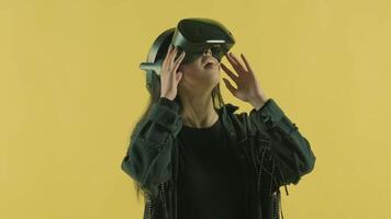 Young woman looks around and wonders how amazing in the VR world. Virtual reality helmet on the yellow background. Portrait video