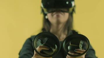 Young woman playing with controllers and wonders how amazing in the VR headset. Virtual reality helmet on the yellow background. Portrait video