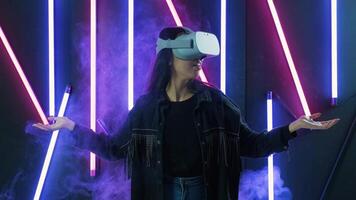 Young woman looks around and wonders how amazing in the VR headset. Virtual reality helmet on color lighting smoke background video