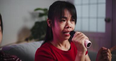 Handheld selective focus shot, Two Asian women sitting on a sofa happily singing karaoke with smiles and enjoying a fun time together at home. Share good times video