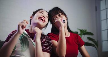 Handheld low angle view shot, Two Asian women sitting on a sofa happily singing karaoke with smiles and enjoying a fun time together at home. Share good times video