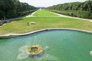 Stunning view of the symmetrical garden with a round fountain with clear water, a long green lawn and neatly trimmed trees. photo
