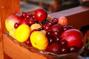 Bright summer abundance. A colorful arrangement of fresh fruits including lemons, apples, cherries, plums and apricots, neatly arranged in a glass vase on a wooden deck. photo