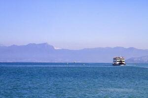 A ferry boat sails through the calm waters of Lake Garda, Italy, against a backdrop of mountains and clear blue sky. photo