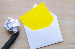 Top view of empty yellow notepad inside an envelope with crumpled paper and pen with customizable space for text photo