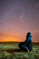 Side view of a man sitting on the grass wearing a jacket and hat, looking up at the milky way, using a headlamp photo