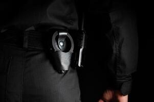 Security guard stands with his back to the camera. Policeman in black uniform with handcuffs and radio on his belt, close-up on black background photo