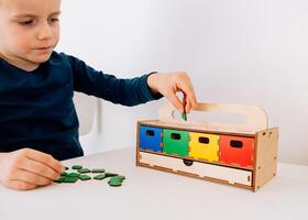 A little boy of 4 years old is arranging multicolored geometrical figures in corresponding boxes. red, yellow, green and blue. Green-colored geometric shapes are scattered on the table. This photo