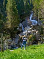 A girl stands near a waterfall surrounded by lush trees. The water cascades down the rocks, creating a natural and serene atmosphere photo