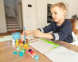 boy takes acrylic paint with a brush from a can in front of him a sheet of paper, a boy of European appearance 4-5 years old paints with paints at home photo