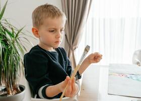a boy of European appearance 4-5 years old sits at a table with an art brush in his hands and thoughtfully thinks what to draw in front of him paints and a sheet of paper photo