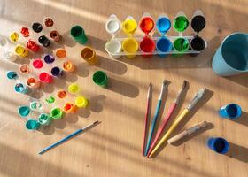 A set of finger paints, gouache, and brushes is arranged on a wooden table. Natural sunlight from the side casts soft shadows, emphasizing the artistic tools and vibrant colors photo