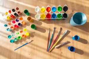 Finger paints and gouache with brushes are scattered on a wooden table. Sunlight from the side highlights the textures and colors, creating a warm and creative atmosphere photo