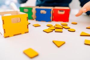 The little pupil arranges multicolored geometric shapes into the corresponding boxes. red, yellow, green and blue. yellow geometric shapes are scattered on the table. This Montessori-inspired photo