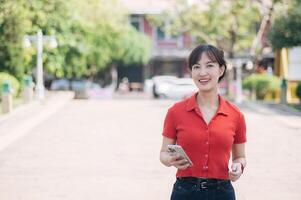 Asian cheerful woman in red shirt holding smartphone and earbuds, standing outdoors on sunny day with blurred background of trees and buildings photo